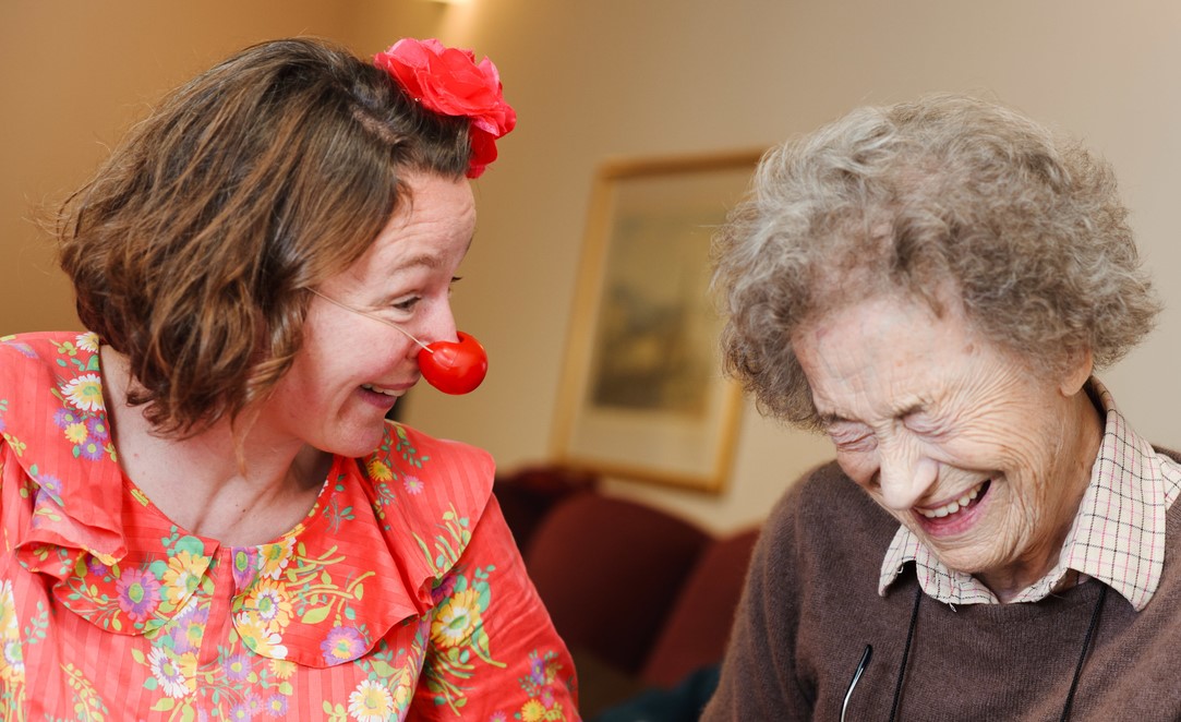 Woman dressed as a clown making an older person laugh.